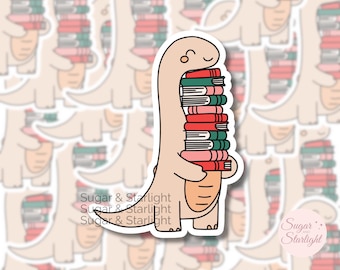 Puede incluir: Una pegatina con un dinosaurio marrón claro sonriente que lleva una pila de libros. Los libros son de color rojo, rosa y verde. La pegatina tiene un borde blanco y el texto "Sugar & Starlight".