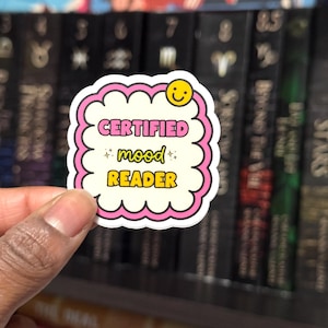 Op de afbeelding: Een witte sticker met een roze rand en de tekst "CERTIFIED mood READER" in roze en geel. De sticker heeft een smiley en wordt voor een boekenplank gehouden.