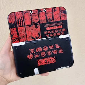 May include: Black and red Nintendo 3DS handheld game console. The top cover features a red and black graphic design with the word "SUPERNOVA". The bottom half has the text "ONE PIECE" and various character emblems.