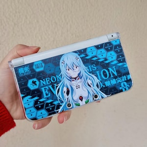 May include: A handheld gaming device with a blue and white design featuring an anime character. The device has a blue background with the words "NEON EV IS ION" and Japanese text. The character has blue hair and is wearing a white and red outfit.