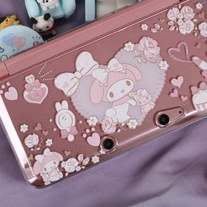Può includere: Una console di gioco portatile rosa con un design decorativo con un personaggio dei cartoni animati, cuori e accenti floreali. La console ha una finitura lucida ed è circondata da altri oggetti decorativi.