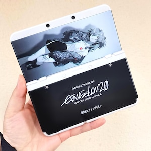 May include: A white and black Nintendo 3DS handheld game console with Evangelion anime artwork. The top screen shows a character illustration, and the bottom features the text "GROUNDWORK OF EVANGELION 2.0" and Japanese text.