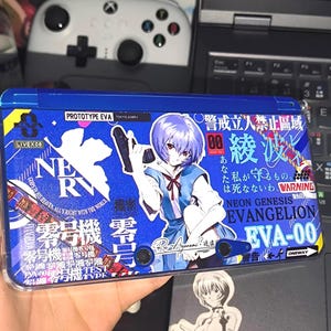 May include: Blue handheld gaming device with an anime-style graphic featuring a character with purple hair and Japanese text. The device has a blue and white design with the text "NE RV" and "EVANGELION".