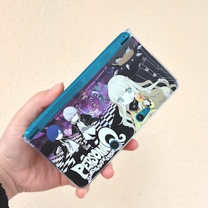 May include: A teal Nintendo 3DS handheld game console featuring a graphic design from the Persona series. The design includes anime-style characters, the word "Persona," and the phrase "Shadow of the Labyrinth" in white lettering. The console is held in a hand.