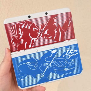 May include: Two Nintendo 3DS consoles, one red and one blue, are stacked. The red console features a red and white design with a graphic of a creature. The blue console has a blue and white design with a different creature graphic.