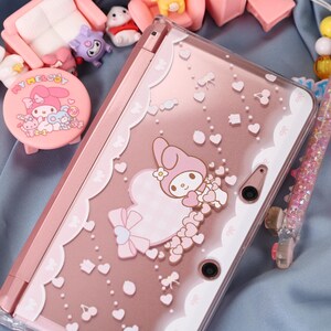 May include: Pink and clear Nintendo 3DS case featuring a My Melody design with hearts and bows. The case has a clear, decorative border and is surrounded by other pink accessories.