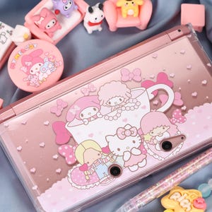 May include: Pink Nintendo 3DS console with a clear cover featuring cartoon characters, bows, and hearts. The console is surrounded by small pink figurines and accessories, including a My Melody compact mirror and a beaded bracelet.