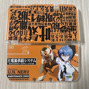 May include: An orange phone case featuring Japanese text and anime-style illustrations. The case has a clear plastic cover and displays images of characters and text, including "MAIN ENERGY SUPPLY SYSTEM" and "U.N. NERV MAINTENANCE CREW".