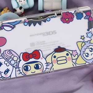 May include: Clear Nintendo 3DS case with colorful cartoon character designs. The case features a white background with various characters in pink, blue, yellow, and red. The text "NINTENDO 3DS" is visible on the case.