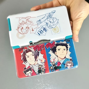 May include: A handheld gaming console with a white top and red and blue bottom. The top features cartoon illustrations in red and blue. The bottom displays two anime-style characters with Japanese text. The console is held by a hand.