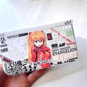 May include: White handheld gaming console with a colorful graphic featuring an anime character in a red outfit. The console has Japanese text and the words "Neon Genesis Evangelion" and "EVA-02".