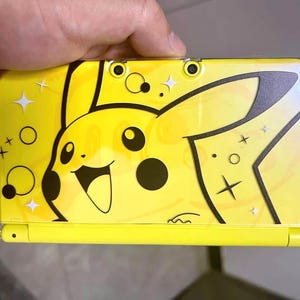 Puede incluir: Consola de juegos portátil amarilla con un diseño de Pikachu. La consola es de color amarillo brillante con detalles en negro y patrones de estrellas y círculos. La consola está sujeta por una mano.