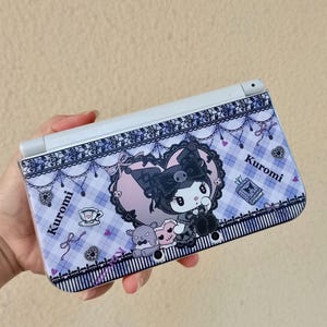 May include: A handheld gaming console featuring a Kuromi design. The console has a white frame and a cover decorated with a cartoon Kuromi character, hearts, and the word "Kuromi" in a decorative font. The background is a light purple plaid pattern.