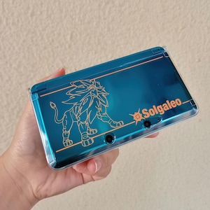 Op de afbeelding: Een teal Nintendo 3DS handheld gameconsole met een Solgaleo-ontwerp. Het ontwerp toont een gouden omtrek van een leeuwachtig wezen en het woord "Solgaleo". De console wordt in de hand van een persoon gehouden.