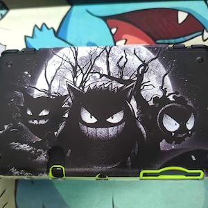 May include: Black and white Nintendo 2DS XL case featuring a spooky design with Gengar, Haunter, and Gastly in front of a full moon and bare trees. The case has neon green accents.