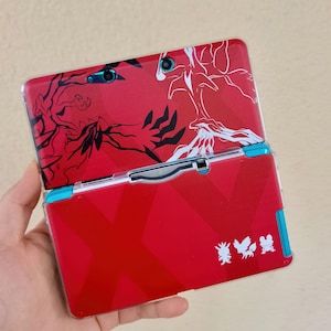 May include: Red handheld gaming console with a decorative cover featuring black and white abstract designs. The console has a blue accent on the side and a series of small character graphics.