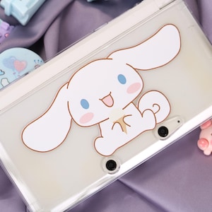 May include: A white handheld gaming console with a clear protective case featuring a cartoon character with large ears, blue eyes, and pink cheeks. The console is on a purple surface.