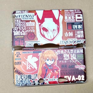 May include: A two-piece Nintendo 3DS case featuring artwork from Neon Genesis Evangelion. The top piece has a red and white design with the text "EVANGELION" and a character illustration. The bottom piece features Asuka Langley Soryu and the text "EVA-02".