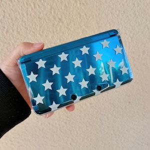 May include: A blue handheld gaming console with a white star pattern. The console is held in a clear protective case. The device has two small camera lenses and a hinge.