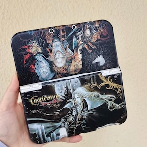 May include: A black Nintendo 3DS case featuring artwork from the video game "Castlevania: Harmony of Dissonance." The case is open, revealing detailed illustrations of characters, a full moon, and the game's title. The case is held in a hand.