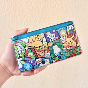 Op de afbeelding: Een teal en veelkleurige Nintendo 3DS-hoes met verschillende Pokémon-personages zoals Pikachu, Eevee en Snorlax. De hoes heeft een glanzende afwerking en een scharnierend ontwerp, met de Pokémon-personages in stripboekstijl gerangschikt.