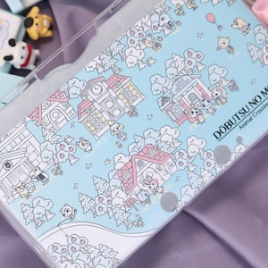 May include: A light blue phone case with a cartoon village design, including houses, trees, and characters. The case has a clear border and the text "DOBUTSU NO MORI Animal Crossing".