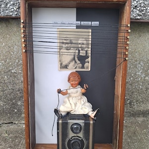 Puede incluir: Una obra de arte enmarcada de técnica mixta con una muñeca vintage sentada sobre una cámara negra. La obra incluye una fotografía en blanco y negro de dos bebés, con el texto "may they have love beside them". El marco es marrón.