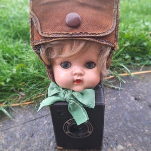 May include: A vintage doll with blue eyes, wearing a brown hat and a green bow tie, is placed on top of a black camera. The doll's face is visible, with blonde hair and a red painted mouth. The camera has a circular lens.