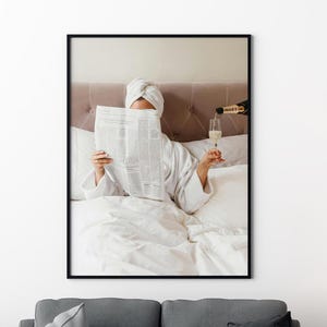Puede incluir: Una impresión enmarcada muestra a una persona con una bata y toalla blancas leyendo un periódico en la cama. Se ve una copa de champán y una botella. La impresión cuelga sobre un sofá gris, una mesa de centro de madera y una lámpara de pie.