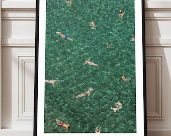 May include: A framed art print featuring an aerial view of people swimming in clear turquoise water. The black framed print is leaning against a white wall and a wooden floor. The swimmers are scattered throughout the water.