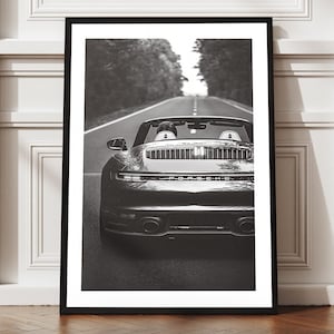 May include: Black and white framed print of a sports car driving on a road. The car is centered in the frame, with the road leading into the distance. The print is displayed against a white wall with decorative molding.