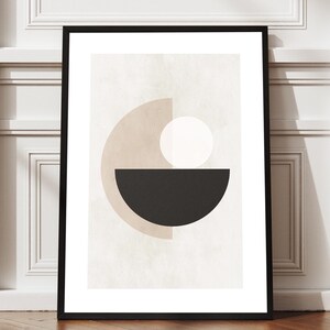 May include: Framed abstract art print featuring geometric shapes in beige, black, and white. The artwork includes a black semi-circle, a beige semi-circle, and a white circle. The print is displayed in a black frame against a white wall and a wooden floor.