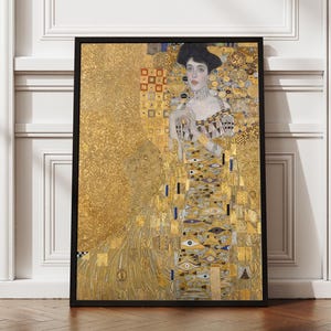 May include: Framed art print of a Gustav Klimt painting. The artwork features a woman in a golden dress with intricate patterns and a black hat. The background is a textured gold, and the frame is black. The print is displayed against a white wall.