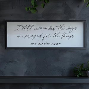 May include: A framed sign with the text "I still remember the days we prayed for the things we have now" hangs above a shelf with potted plants. The sign has a black frame and white background. The plants include succulents and leafy green plants.