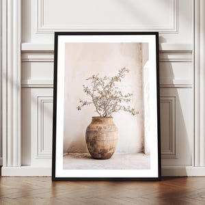 May include: A framed print featuring a large, aged terracotta vase filled with dried branches. The vase is set against a neutral-toned wall, with a glimpse of a window and architectural details. The frame is black, and the floor is a dark wood.