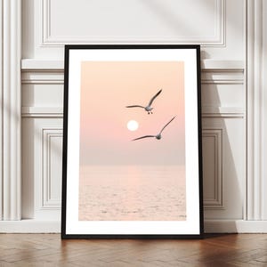 Pode incluir: Impressão artística emoldurada com duas gaivotas em voo contra um céu de pôr do sol pastel sobre o oceano. A obra de arte é exibida em uma moldura preta e repousa sobre um piso de madeira, encostada em uma parede branca.