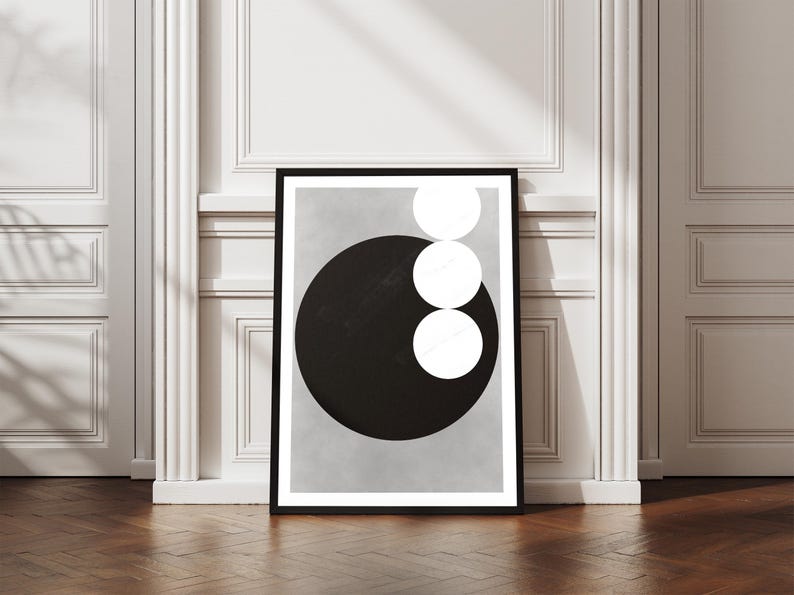 Op de afbeelding: Een ingelijste abstracte kunstprint met een groot zwart cirkel met drie witte cirkels verticaal gestapeld op een lichtgrijze achtergrond. Het kunstwerk wordt getoond tegen een witte muur en een houten vloer.