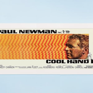 May include: A framed movie poster for "Cool Hand Luke" featuring Paul Newman. The poster has a white frame and a design with orange and red stripes, a profile of Newman, and the film's title.