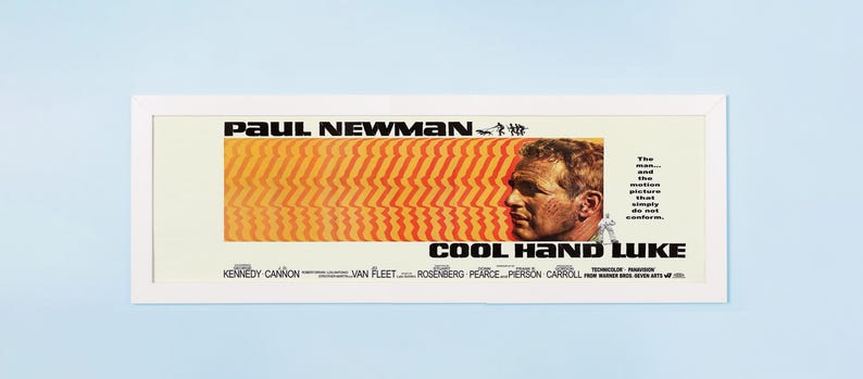 May include: A framed movie poster for "Cool Hand Luke" featuring Paul Newman. The poster has a white frame and a design with orange and red stripes, a profile of Newman, and the film's title.