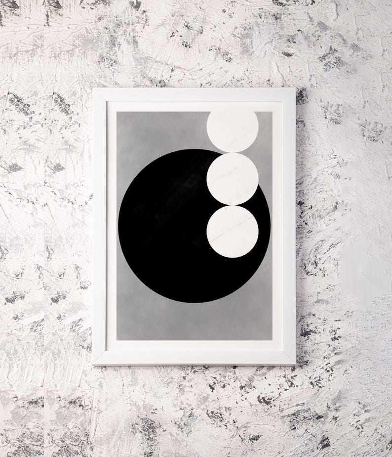 Op de afbeelding: Abstracte kunstafdruk in een witte lijst. Het kunstwerk toont een grote zwarte cirkel met drie witte cirkels die bovenop elkaar zijn gestapeld. De achtergrond is lichtgrijs.