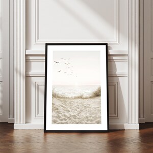 May include: Framed art print featuring a serene beach scene. The image shows a sandy beach with dune grass, the ocean, and a flock of birds in the sky. The print is in a black frame and is displayed against a white wall.