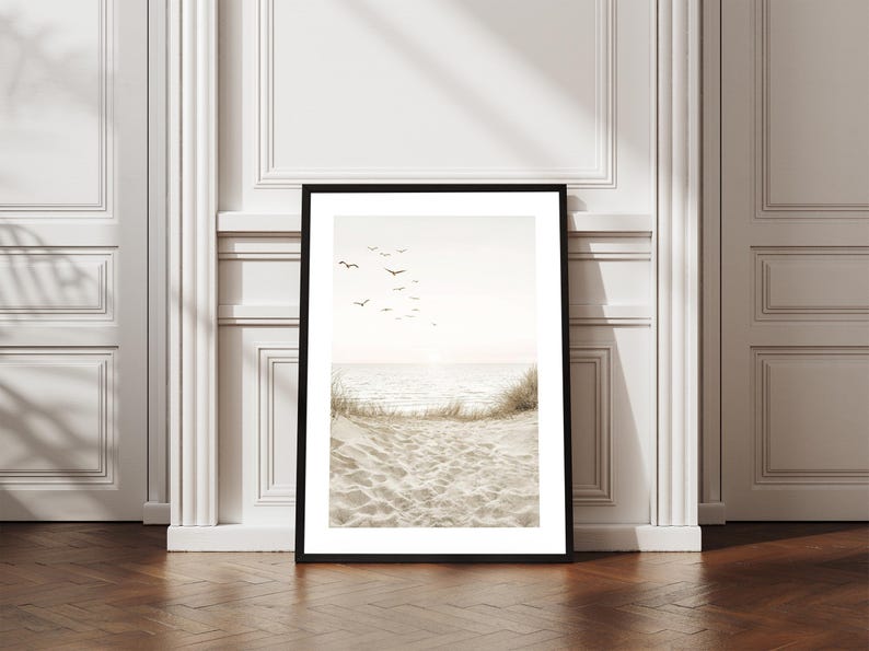 May include: Framed art print featuring a serene beach scene. The image shows a sandy beach with dune grass, the ocean, and a flock of birds in the sky. The print is in a black frame and is displayed against a white wall.