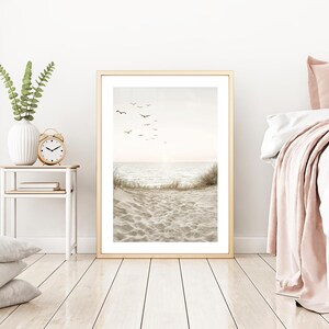 May include: A framed beach scene print with a golden frame. The artwork depicts a serene ocean view with birds in the sky, sand, and waves. The room also features a white vase with greenery, a gold alarm clock, and a bed with pink accents.