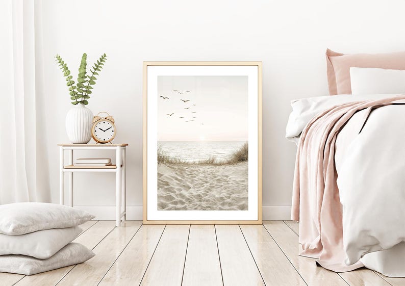 May include: A framed beach scene print with a golden frame. The artwork depicts a serene ocean view with birds in the sky, sand, and waves. The room also features a white vase with greenery, a gold alarm clock, and a bed with pink accents.