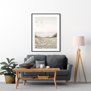 May include: A framed beach scene print with a black frame. The artwork depicts a sandy beach, ocean waves, and birds in flight against a soft, neutral-toned sky. The print is displayed above a gray sofa.
