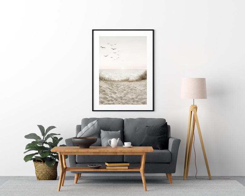 May include: A framed beach scene print with a black frame. The artwork depicts a sandy beach, ocean waves, and birds in flight against a soft, neutral-toned sky. The print is displayed above a gray sofa.