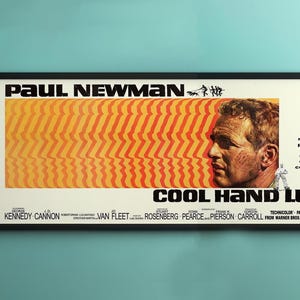 May include: Framed movie poster for "Cool Hand Luke" featuring Paul Newman. The poster has a white background with orange and yellow stripes, a portrait of Newman, and the film's title in bold black letters.
