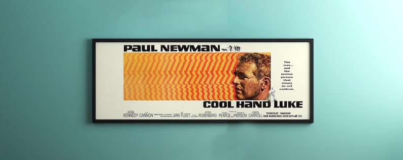 May include: Framed movie poster for "Cool Hand Luke" featuring Paul Newman. The poster has a white background with orange and yellow stripes, a portrait of Newman, and the film's title in bold black letters.