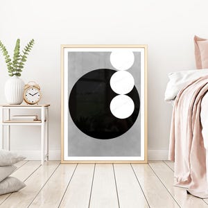 Op de afbeelding: Een ingelijste abstracte kunstprint met een grote zwarte cirkel en drie witte cirkels op een grijze achtergrond. Het kunstwerk wordt tentoongesteld in een slaapkamer met een bed, kussens en een bijzettafel met een vaas en klok.