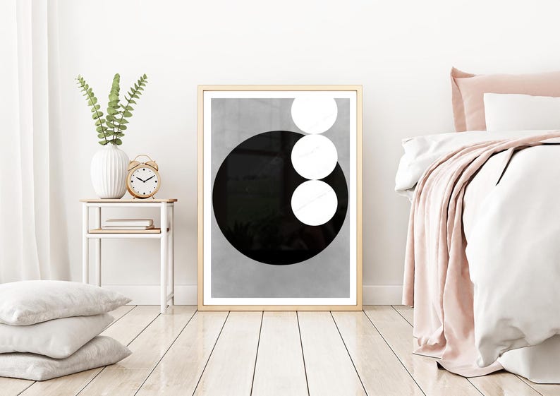 Op de afbeelding: Een ingelijste abstracte kunstprint met een grote zwarte cirkel en drie witte cirkels op een grijze achtergrond. Het kunstwerk wordt tentoongesteld in een slaapkamer met een bed, kussens en een bijzettafel met een vaas en klok.
